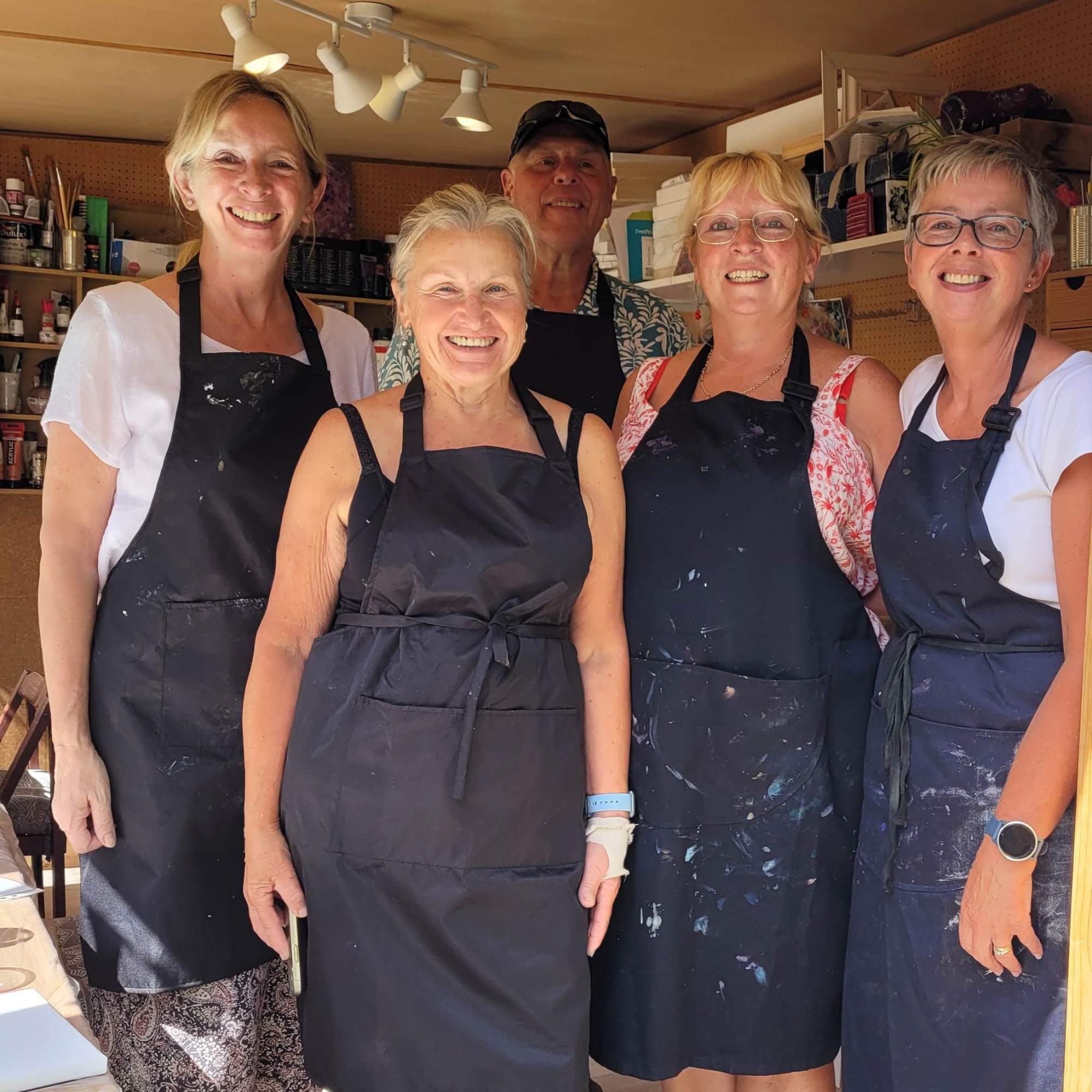 Group of five smiling participants wearing navy aprons in MarchMadeArt creative workshop studio surrounded by art supplies